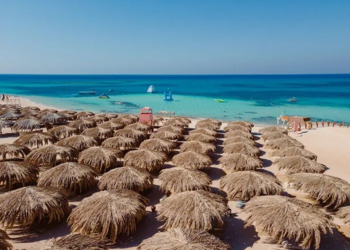 Rows of palm-thatch umbrellas on the beach of Paradise Island with boats in the turquoise lagoon, Hurghada