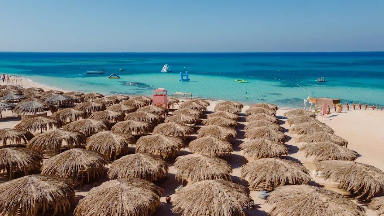 Rows of palm-thatch umbrellas on the beach of Paradise Island with boats in the turquoise lagoon, Hurghada