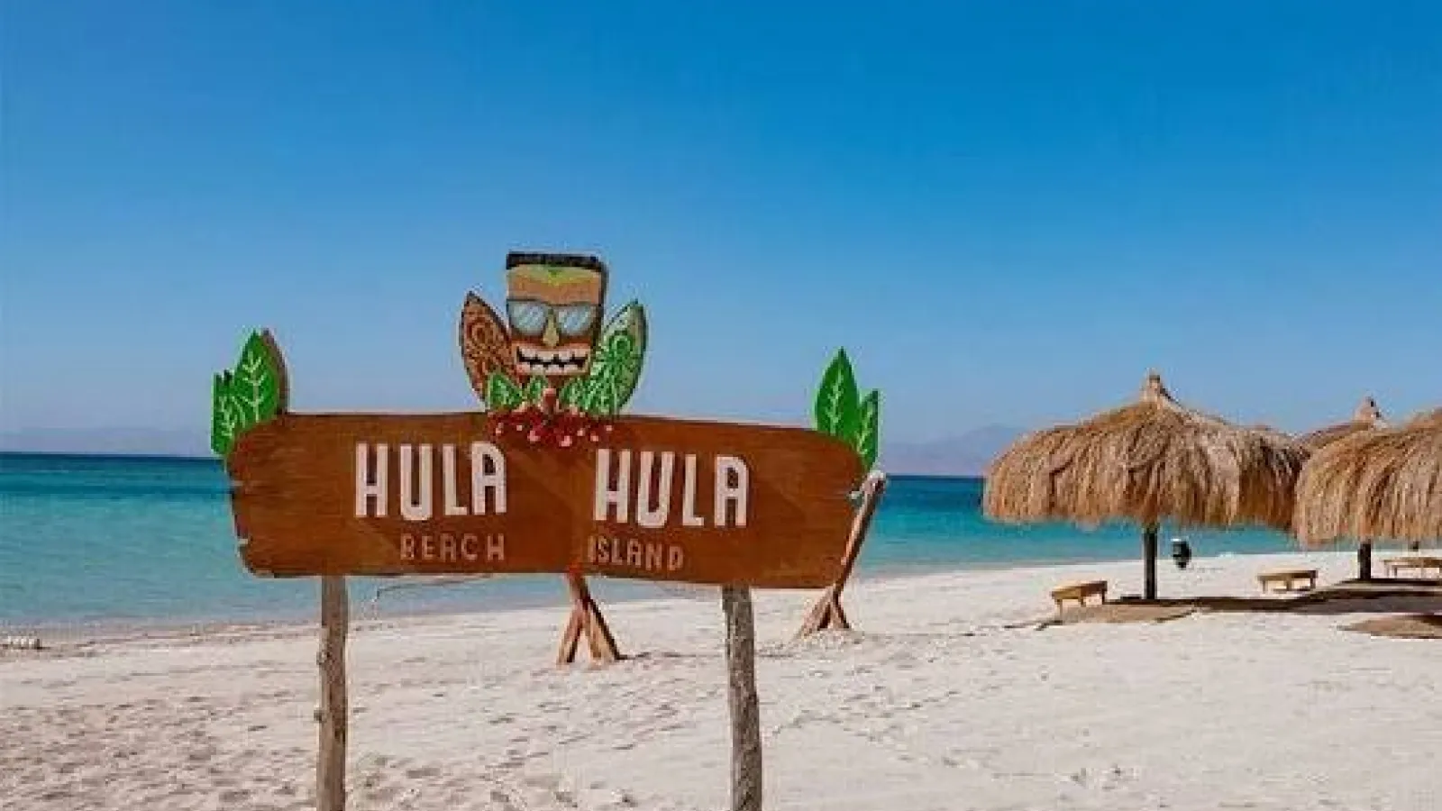 Wooden Hula Hula Beach Island sign on the sand beside thatched umbrellas, Hurghada
