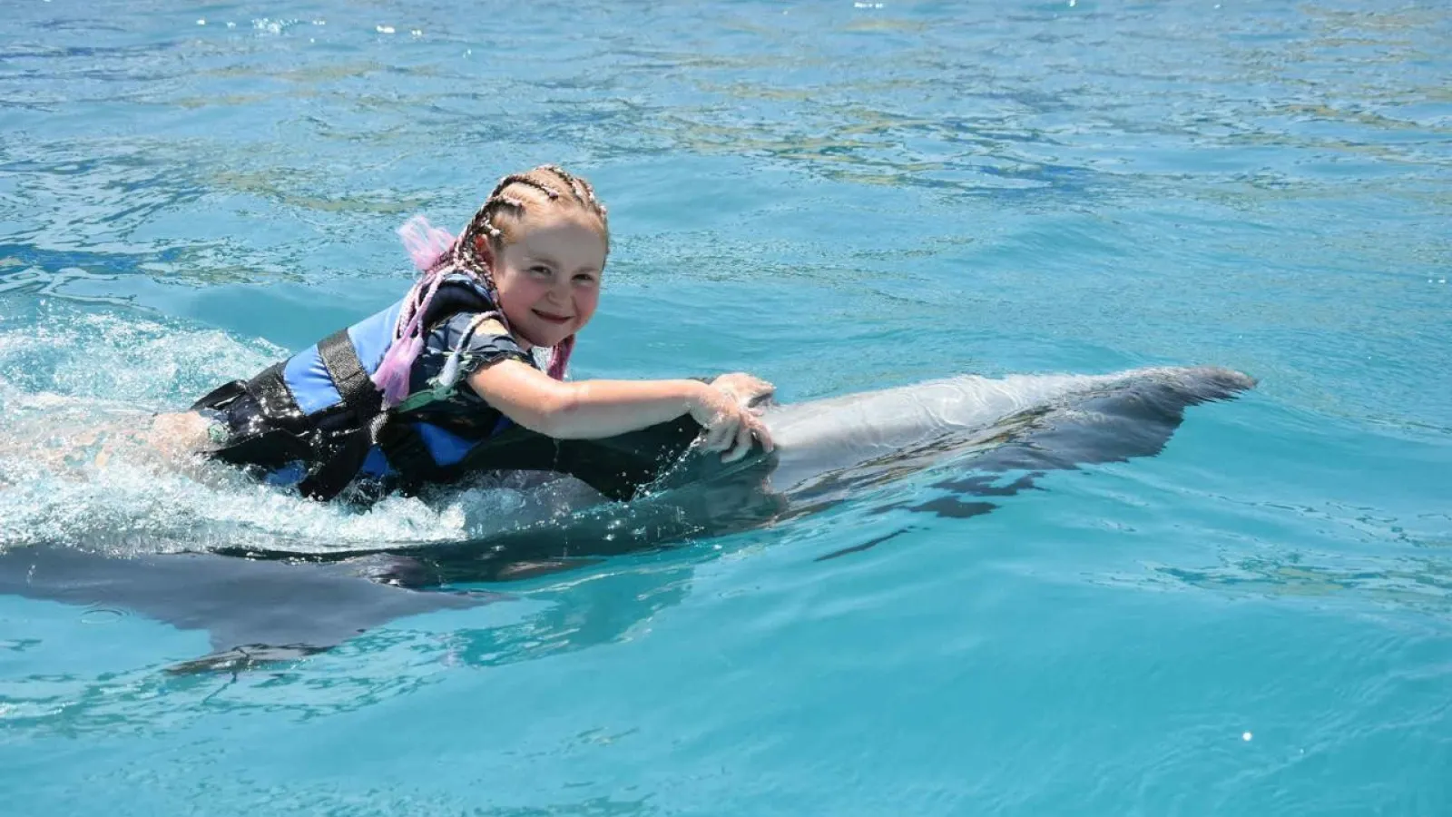 Girl in a life vest holding onto a dolphin while swimming in the dolphinarium pool
